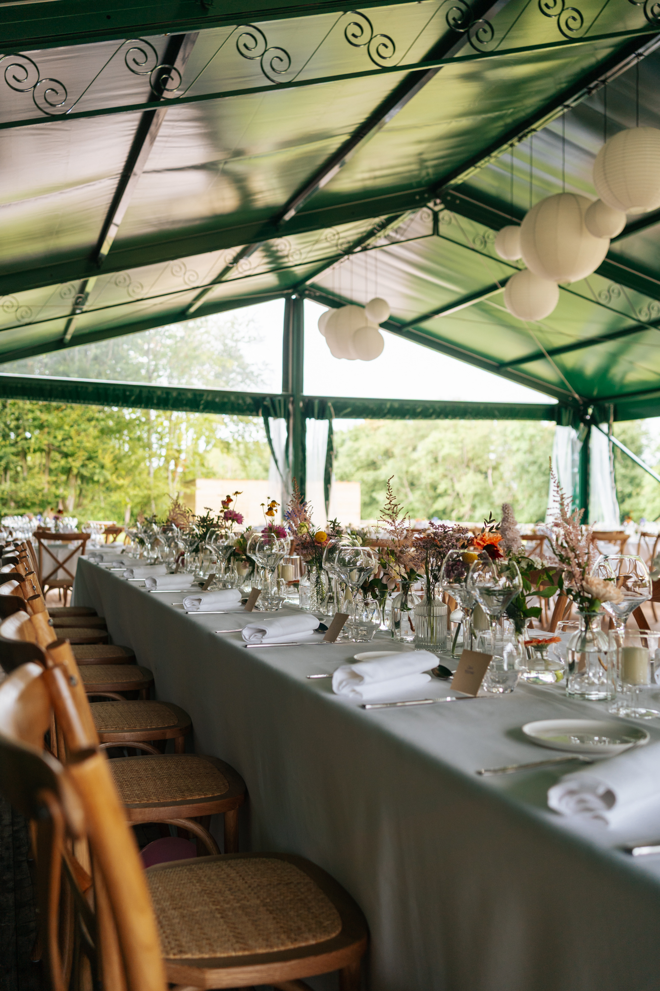 Elegant wedding reception setup under a green marquee, with flower arrangements and wine glasses set on the tables.