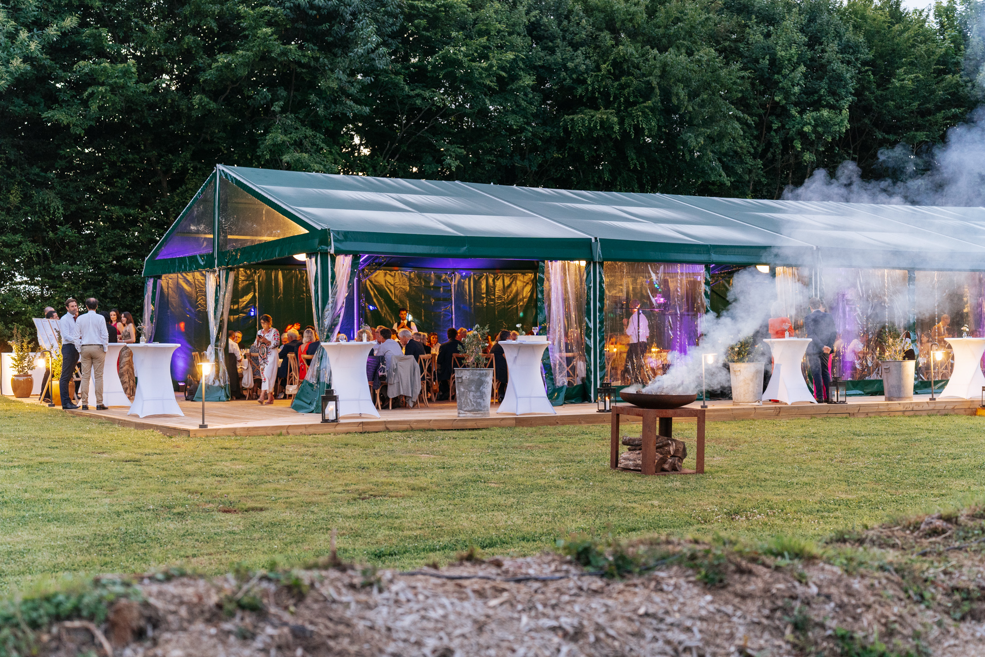 Evening reception in a green marquee with guests enjoying the party, smoke effects and ambient lighting at Domaine de Barneville.